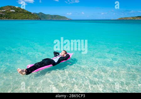 Uomo rilassato in tuta che galleggia su una zattera a bordo piscina rosa brillante in acque blu tropicali Foto Stock