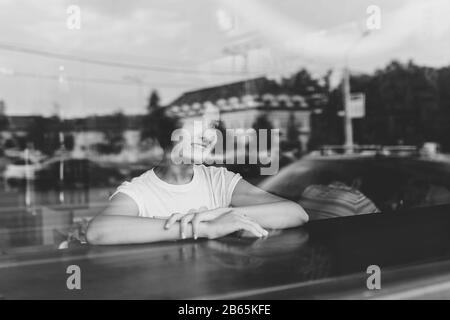 Attraente giovane donna seduto al tavolo in caffetteria. Fotografato dalla strada con riflessi della città nella finestra Foto Stock