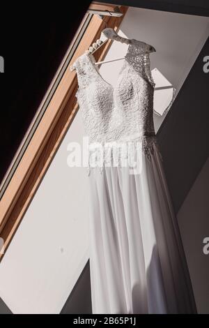 Abito da sposa bianco appende su una spalla vicino alla finestra, prima della cerimonia Foto Stock