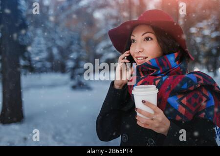 Donna vestita alla moda con un cappotto e un cappello a testa larga in inverno Park bere caffè e parlare sullo smartphone Foto Stock