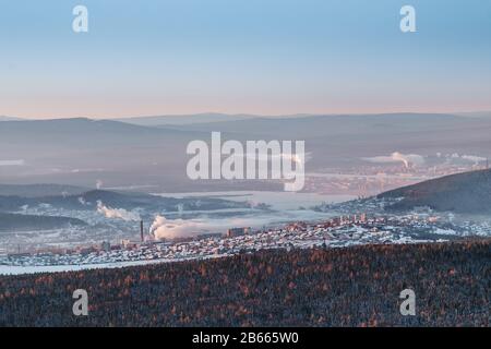 La vista aerea del centro industriale della città degli Urali di Zlatoust con il fumo di tubo dalle fabbriche, circondato da montagne invernali. Foto Stock