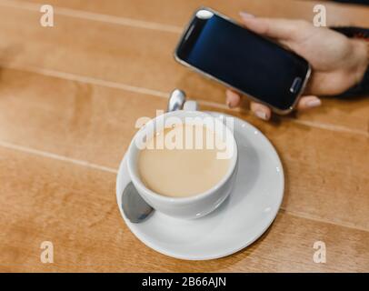 Donna con smartphone che prende tè caldo o caffè con latte in una tazza su sfondo di tavolo di legno. Vista in primo piano Foto Stock