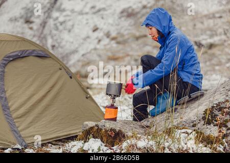 Donna acqua bollente sul campeggio portatile gas-jet vicino alla tenda da trekking in neve montagne tardo autunno Foto Stock