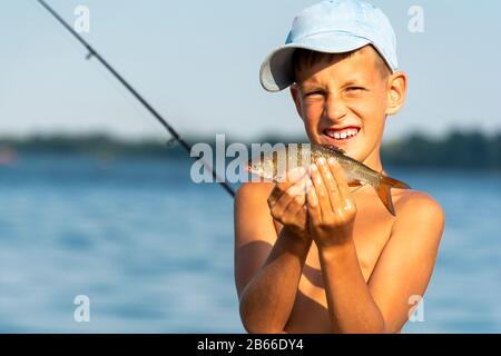 Felice ragazzo sorridente tenendo preso pesce d'acqua dolce in mani contro il fondo di canna e fiume Foto Stock