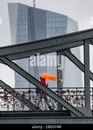 10 marzo 2020, Hessen, Francoforte sul meno: Una donna cammina attraverso il ponte di ferro sotto la pioggia. Dietro di esso si trova la sede della Banca centrale europea (BCE). Presso la BCE vi è un primo caso confermato di coronavirus nel personale. Un membro del team ha testato positivamente il nuovo patogeno, annunciato dalla banca centrale. Foto: Frank Rumpenhorst/Dpa Foto Stock