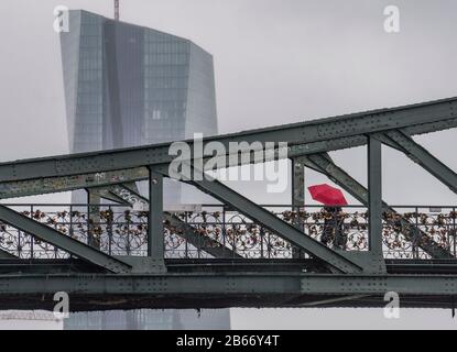 10 marzo 2020, Hessen, Francoforte sul meno: Una donna cammina attraverso il ponte di ferro sotto la pioggia. Dietro di esso si trova la sede della Banca centrale europea (BCE). Presso la BCE vi è un primo caso confermato di coronavirus nel personale. Un membro del team ha testato positivamente il nuovo patogeno, annunciato dalla banca centrale. Foto: Frank Rumpenhorst/Dpa Foto Stock