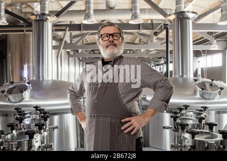 Produttore professionista di birra sulla sua produzione artigianale di alcol. Specialista, uomo anziano in abbigliamento da lavoro che si pone sicuro. Concetto di open business, prodotto ecologico, fabbrica di birra artigianale, fabbrica, fabbrica, avvio. Foto Stock