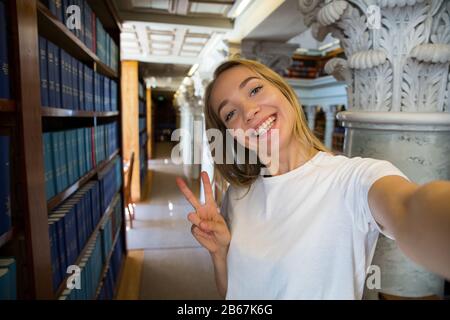 Eccitato giovane ragazza in piedi in vecchia biblioteca tradizionale alle librerie, sorridente e ridente studente facendo selfie sulla fotocamera del telefono, avendo divertimento. Superiore Foto Stock