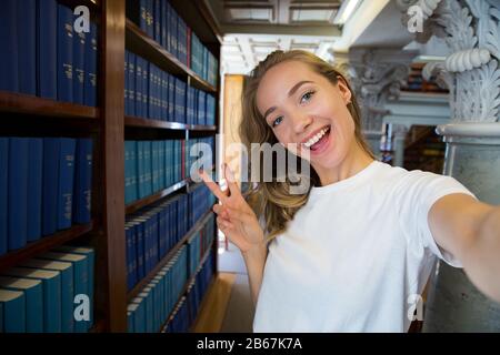 Eccitato giovane ragazza in piedi in vecchia biblioteca tradizionale alle librerie, sorridente e ridente studente facendo selfie sulla fotocamera del telefono, avendo divertimento. Superiore Foto Stock