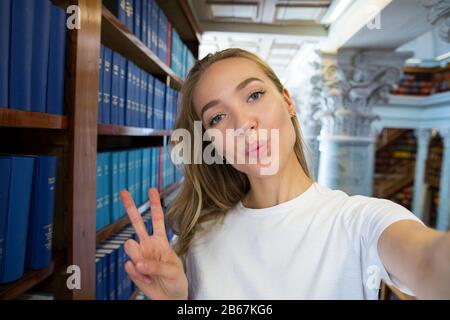 Eccitato giovane ragazza in piedi in vecchia biblioteca tradizionale alle librerie, sorridente e ridente studente facendo selfie sulla fotocamera del telefono, avendo divertimento. Superiore Foto Stock