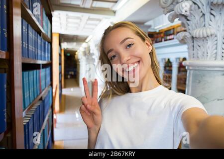 Eccitato giovane ragazza in piedi in vecchia biblioteca tradizionale alle librerie, sorridente e ridente studente facendo selfie sulla fotocamera del telefono, avendo divertimento. Superiore Foto Stock