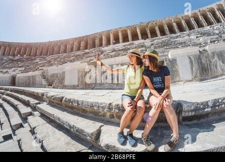 Due giovani ragazze studentesse che viaggiano prendendo selfie l'antico anfiteatro greco Foto Stock