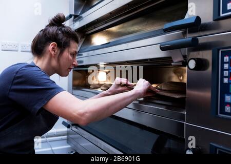 Donna che indossa grembiule in piedi in un panificio artigianale, mettendo pane di pasta di fonte in forno. Foto Stock