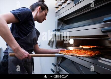 Donna che indossa grembiule in piedi in una panetteria artigianale, mettendo la pizza in forno. Foto Stock