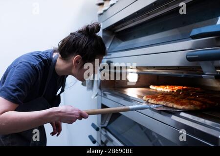 Donna che indossa grembiule in piedi in una panetteria artigianale, mettendo la pizza in forno. Foto Stock