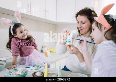 Madre e figlie giovani che celebrano la festa di Pasqua. Ragazza carina piccola con orecchie conigliette che muoiono uovo mentre mamma tenere uovo e altri bambini ragazza guardare Foto Stock