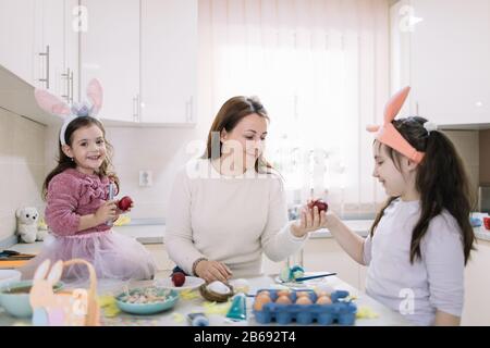 Mamma e figlie che indossano orecchie di coniglio che muoiono uova. Madre sorridente e due sorelle carine che indossano orecchie di coniglio dipingere le uova di Pasqua nella cucina soleggiata. Foto Stock