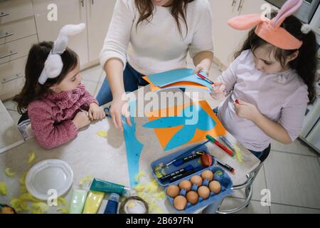 Vista dall'alto delle ragazze al tavolo con uova, vernice e fogli di carta, donna Ritagliata che tiene fogli di carta mentre si siede al tavolo con uova, marcatori, pennelli e Foto Stock