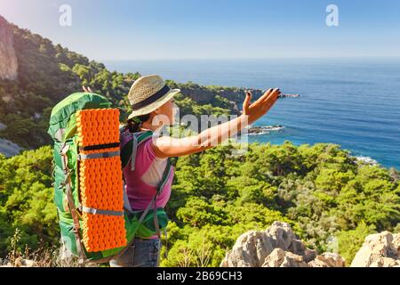 Una giovane donna felice in un viaggio escursionistico con uno zaino sollevato le mani ammirando la colorata vista sul mare. Viaggio attraverso la Via Licia in Turchia Foto Stock