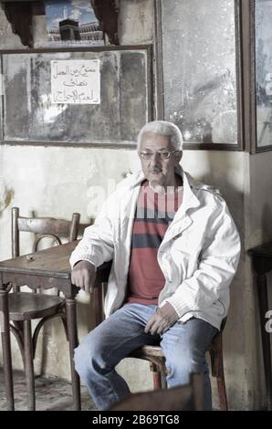 Questo suggestivo ritratto cattura un uomo anziano seduto da solo in un'antica caffetteria egiziana. La sua espressione contemplativa, capelli bianchi e abbigliamento semplice. Foto Stock