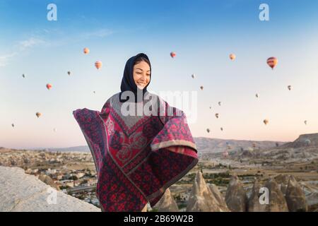 Donna in abiti tradizionali poncho guardando splendida vista di un colorato aria calda palloncini che volano sulla valle a Cappadocia, Turchia. Foto Stock