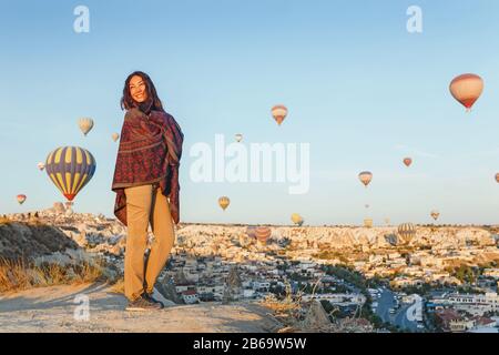 Donna in abiti tradizionali poncho guardando splendida vista di un colorato aria calda palloncini che volano sulla valle a Cappadocia, Turchia. Foto Stock