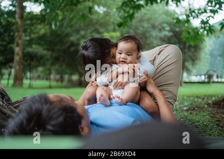 due bambini si rilassano seduti sullo stomaco di un padre quando si distende sull'erba Foto Stock
