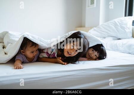 tre bambini giocano e si nascondono sotto le coperte sul letto quando si svegliano Foto Stock