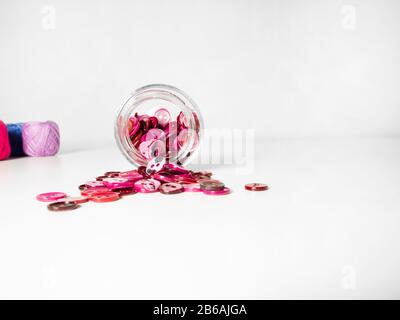 Vaso in vetro pieno di bottoni rosa e fili di abbigliamento su sfondo bianco. Concetto di cucito e di mestiere. Foto Stock