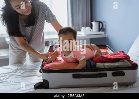 La mamma sorride tenendo un bambino carino sdraiato in una valigia aperta piena di vestiti posti sul letto Foto Stock