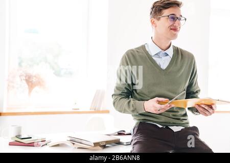 Immagine di un giovane studente ottimista positivo sedersi sul tavolo al coperto in occhiali che tengono appunti di scrittura note. Foto Stock