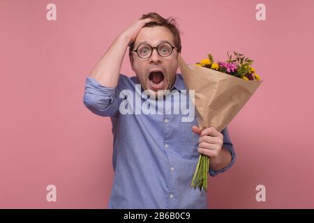 Giovane caucasico in bicchieri che ha sorpreso il viso con fiori per la sua ragazza per il regalo di compleanno. Studio girato su parete rosa Foto Stock