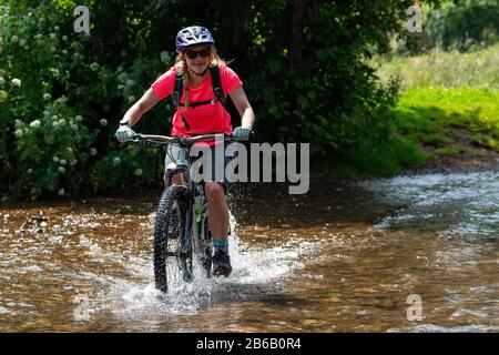 Una giovane donna bionda guida una mountain bike attraverso un ruscello vicino a Minehead nel Somerset. Foto Stock