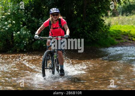 Una giovane donna bionda guida una mountain bike attraverso un ruscello vicino a Minehead nel Somerset. Foto Stock