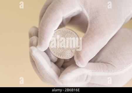 Le mani nei guanti bianchi tengono con attenzione una moneta d'argento bitcoin. Speranza di crescita del capitale Foto Stock