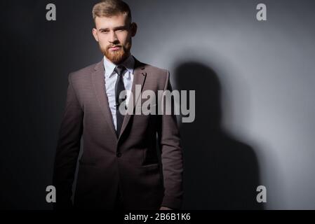 Studio ritratto di un giovane bel ragazzo bearded di venticinque anni, in un vestito ufficiale, guardando la macchina fotografica. Su uno sfondo scuro. Drammatico l Foto Stock