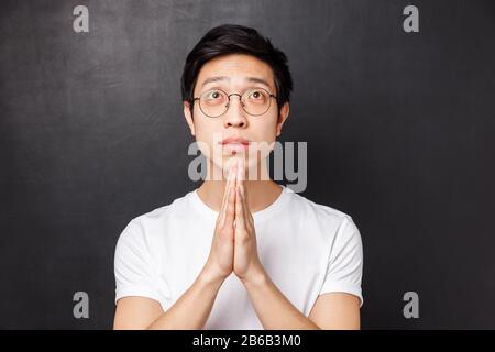 Primo piano ritratto di giovane ragazzo asiatico pieno di speranza in occhiali e t-shirt bianca, tenere le mani insieme in pregare, cercando pleading, anticipando Foto Stock