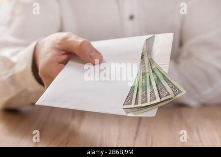 La mano dell'uomo contiene una busta bianca riempita di banconote della valuta polacca e la consegna sopra il tavolo. Foto Stock