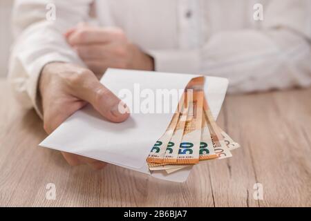 La mano di un uomo tiene una busta bianca riempita di banconote in euro e la consegna sopra il tavolo. Foto Stock