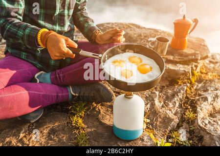 Un viaggiatore donna che cucinava e mangiava le uova strapazzate e il caffè all'alba sulle nebbie montagne. Attrezzature da campeggio e concetto di avventura Foto Stock