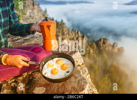 Un viaggiatore donna che cucinava e mangiava le uova strapazzate e il caffè all'alba sulle nebbie montagne. Attrezzature da campeggio e concetto di avventura Foto Stock