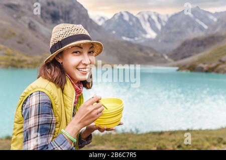 Una donna escursionista mangia il pranzo e gode di una vista sul lago blu in montagna, concetto di cibo da campeggio Foto Stock