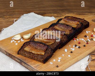 Fette di pancetta e prezzemolo su pane tostato nero, pancetta di maiale con spezie e aglio sono su un supporto scuro. Delizioso sandwich appetitoso con carne del Foto Stock