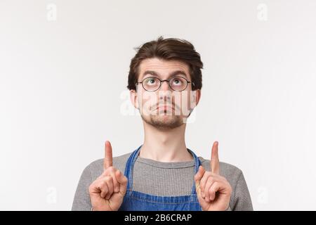 Primo piano ritratto di cupo bello bearded giovane studente maschio iniziare carriera come barista, indossare paron e occhiali, puntando le dita in su e pouting Foto Stock