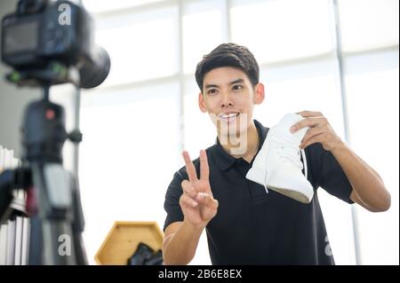 Social media influencer che rivede la moda scarpe. Sorridente giovane uomo che vlogging circa la scarpa sportiva degli uomini e filmarsi a casa su una videocamera. Foto Stock