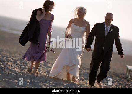 Sorridente sposa e sposo che camminano lungo una spiaggia di sabbia con una delle loro bridesmaids. Foto Stock