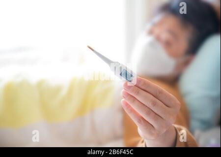 Un uomo asiatico sdraiato a letto, indossando una maschera monouso, misurando la temperatura corporea Foto Stock