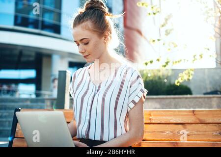 Bella donna d'affari zenzero sta digitando qualcosa al pc mentre si siede sulla panchina Foto Stock