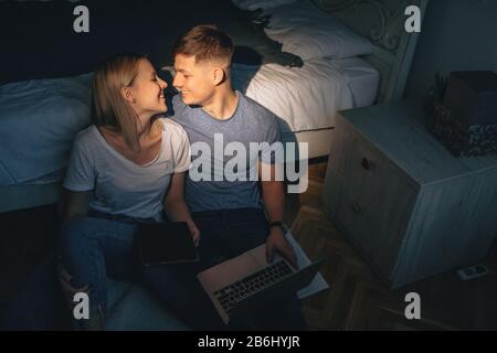 Incantevole coppia caucasica baciare l'un l'altro mentre si siede sul pavimento e utilizzando un tablet e un computer portatile Foto Stock