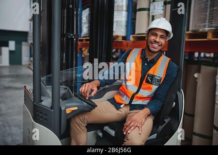 Ritratto del giovane manager africano felice del carrello elevatore a forche indossando giubbotto di sicurezza e hardhat bianco per il trasporto di merci da uno scaffale all'altro mentre si guarda dentro Foto Stock